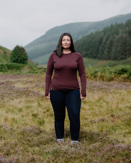 A woman stands in a grassy field with mountains and a forest in the background, wearing a long-sleeve burgundy top and dark navy pants.