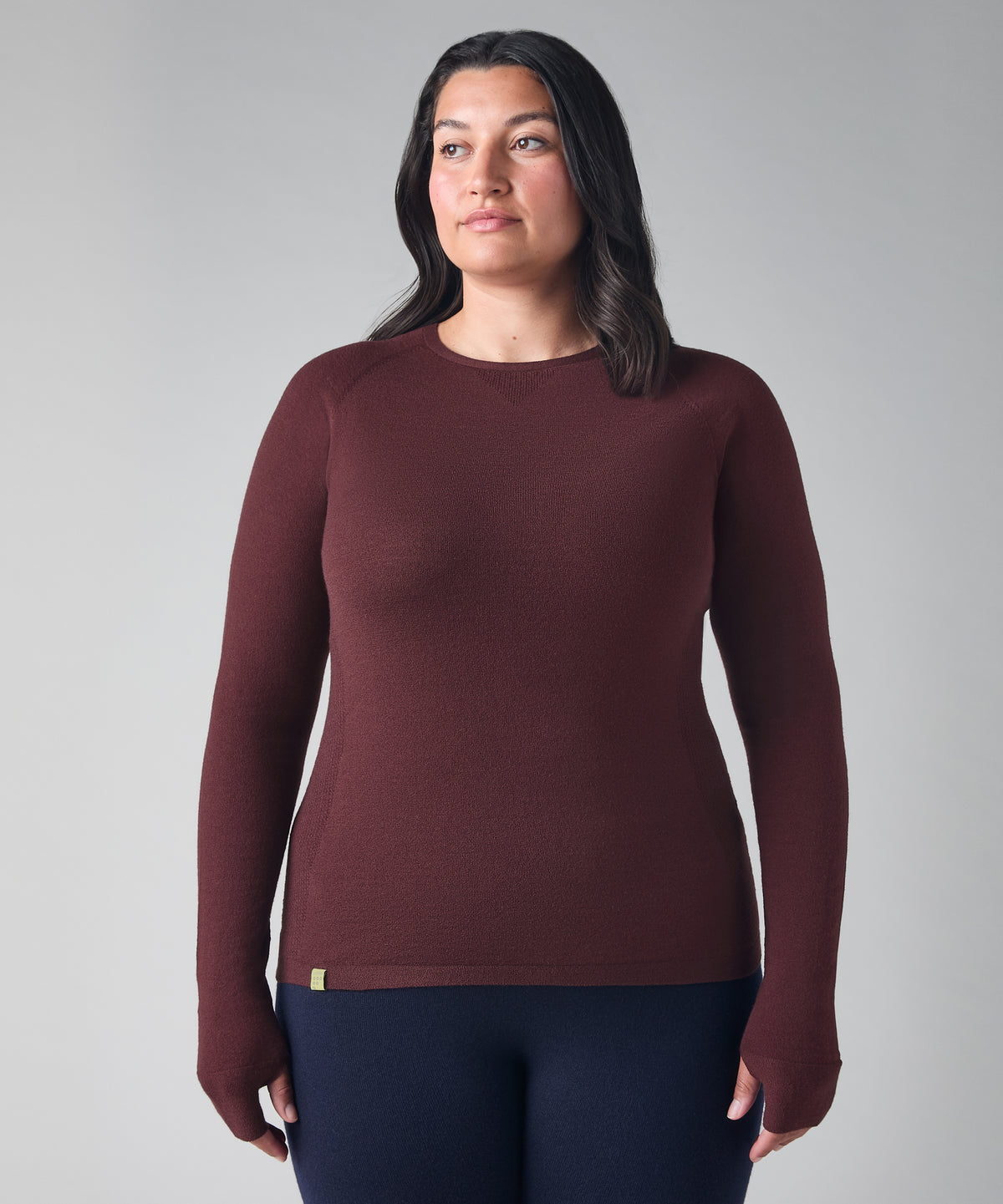 A woman modeling a long-sleeve crewneck knit top in a deep burgundy color, paired with dark navy leggings, against a neutral gray studio background.