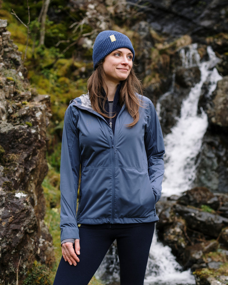 Woman in a blue jacket and beanie standing in front of a waterfall