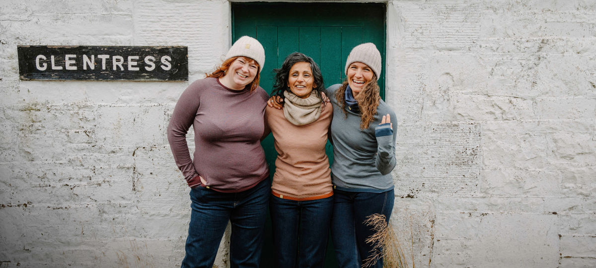 Three women standing together in front of a white textured wall with a green door and a black sign reading 'GLENTRESS'. They are wearing knit beanies and layered tops in muted earthy tones.