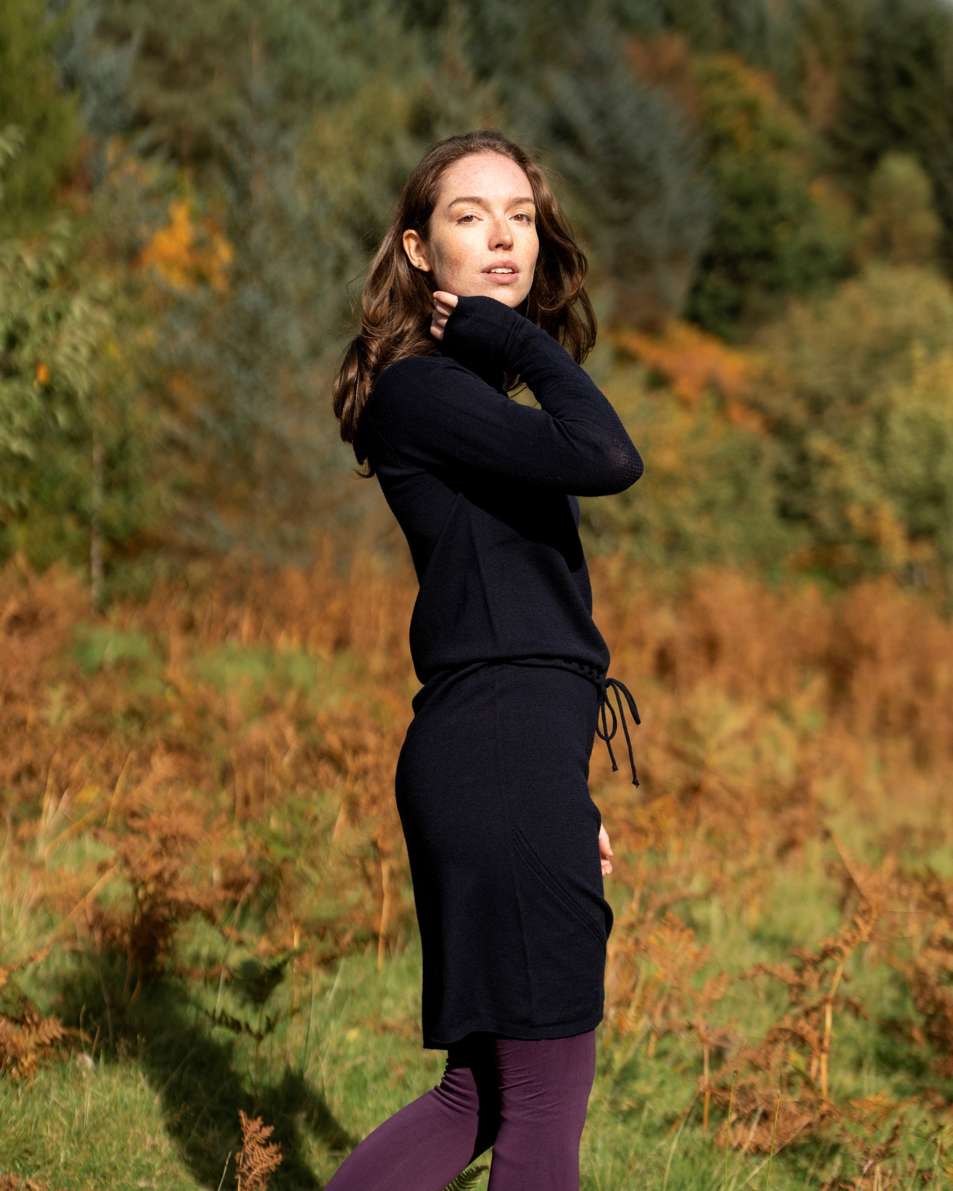 Woman in dark navy merino wool dress standing in a forest with autumn foliage