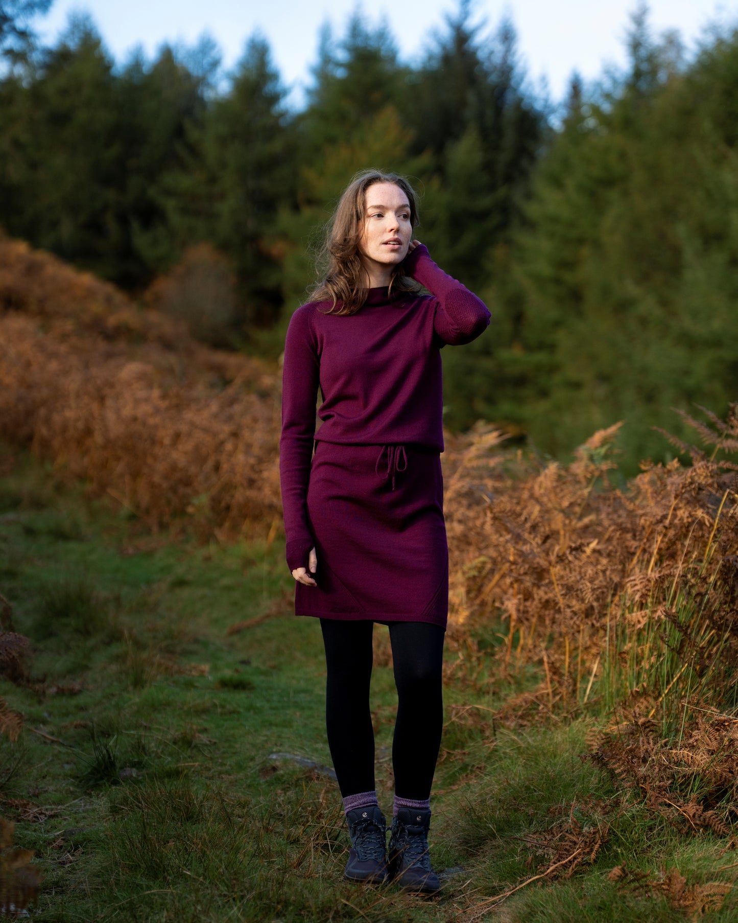 Woman in aeggplant merino wool dress standing in a forest with trees and grass in the background