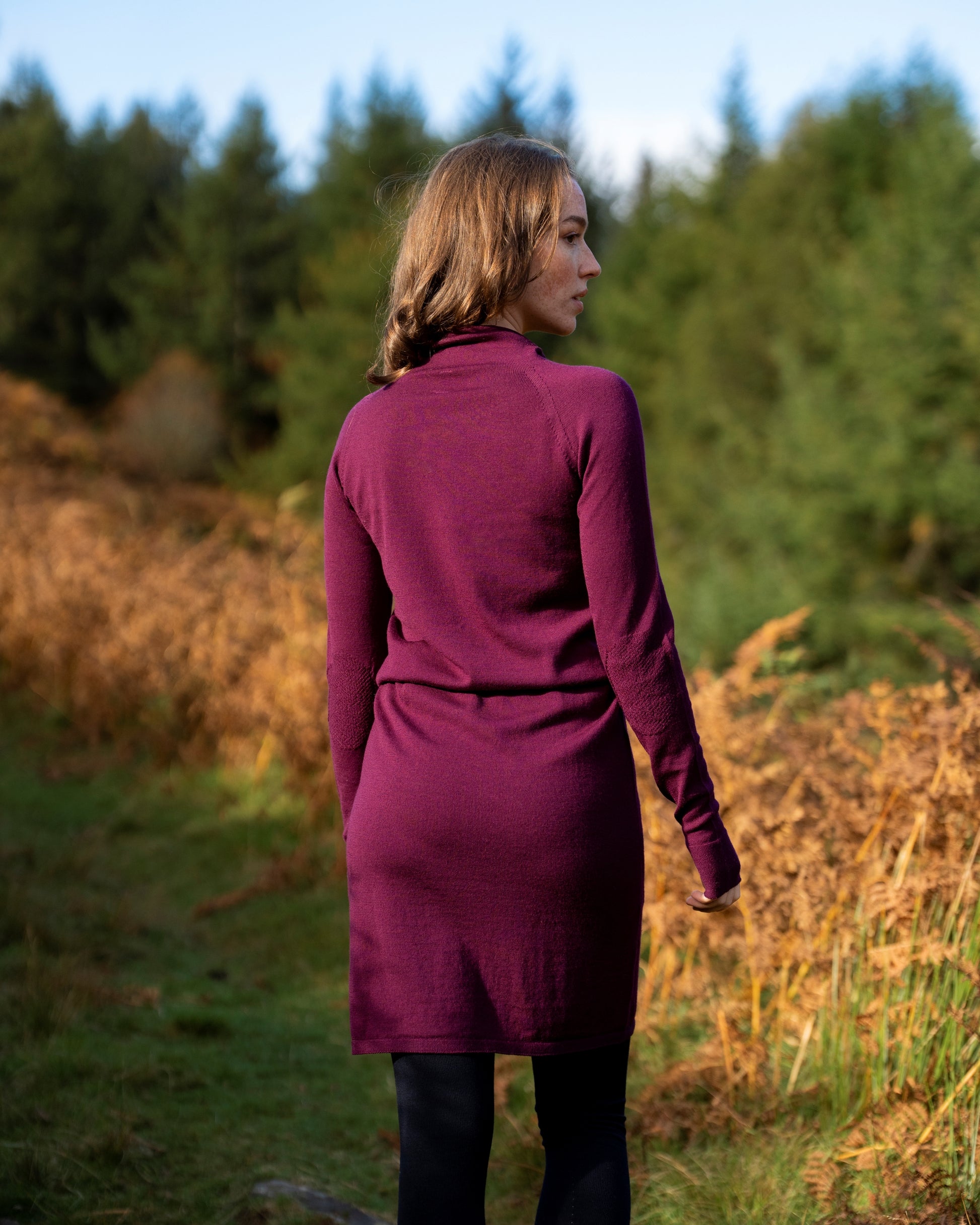 Woman in an eggplant merino wool dress standing in a natural setting with trees and grass.