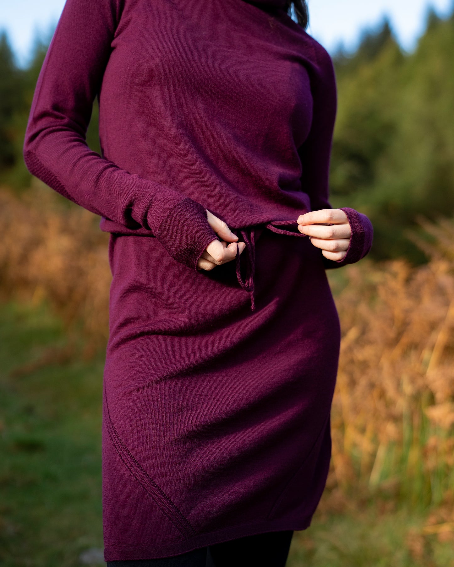 Person wearing an eggplant merino wool dress with tie waist and thumb passes with a blurred natural background