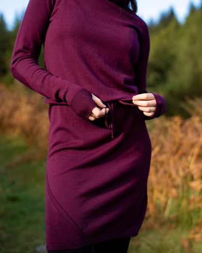 Person wearing an eggplant merino wool dress with tie waist and thumb passes with a blurred natural background