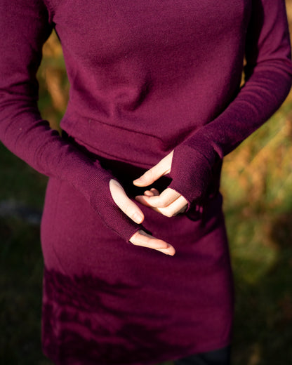 Person wearing a long-sleeve eggplant merino wool dress with thumbpasses in a blurred natural background