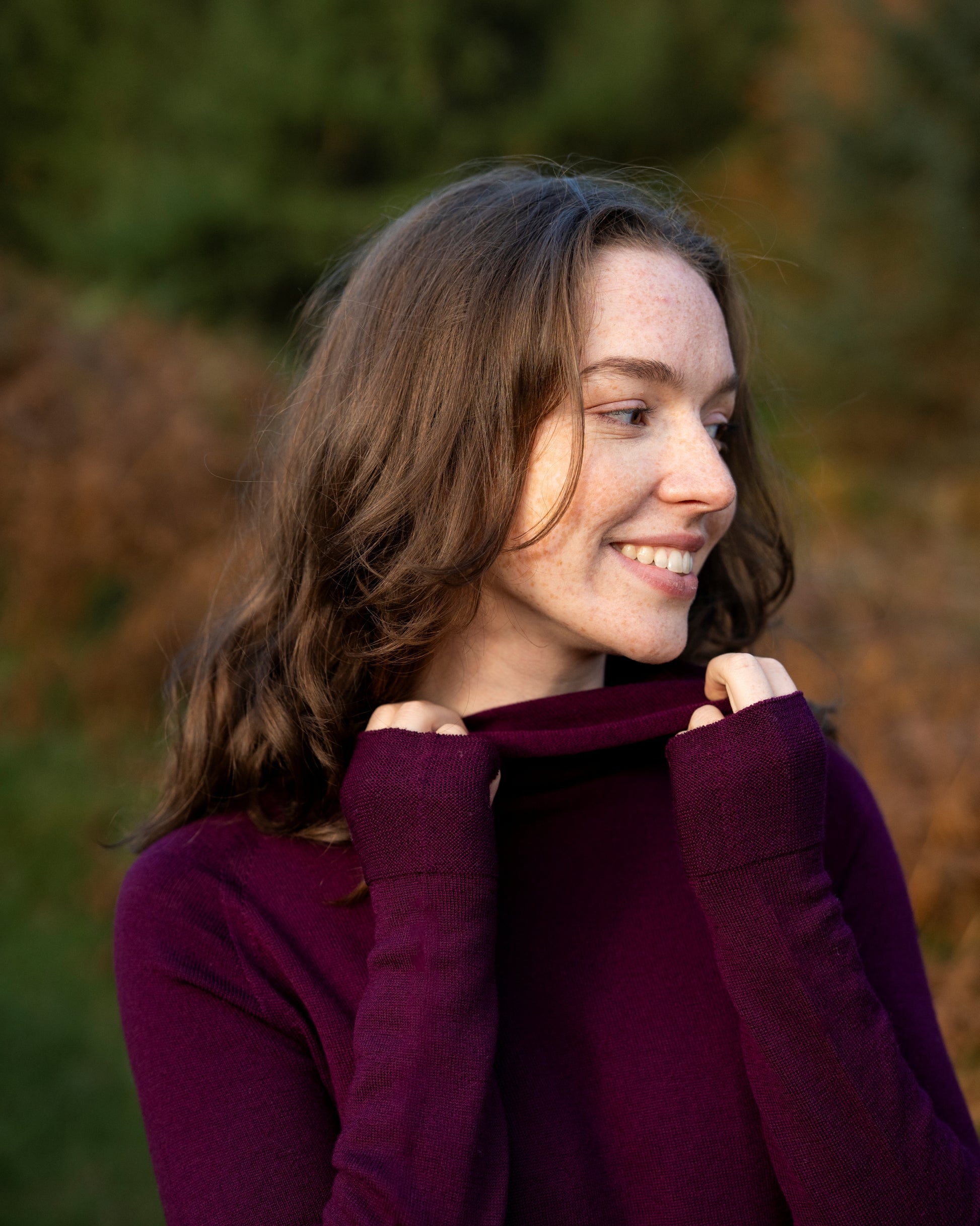 Woman wearing an eggplant merino wool dress with a cowl neck in an outdoor setting with blurred greenery