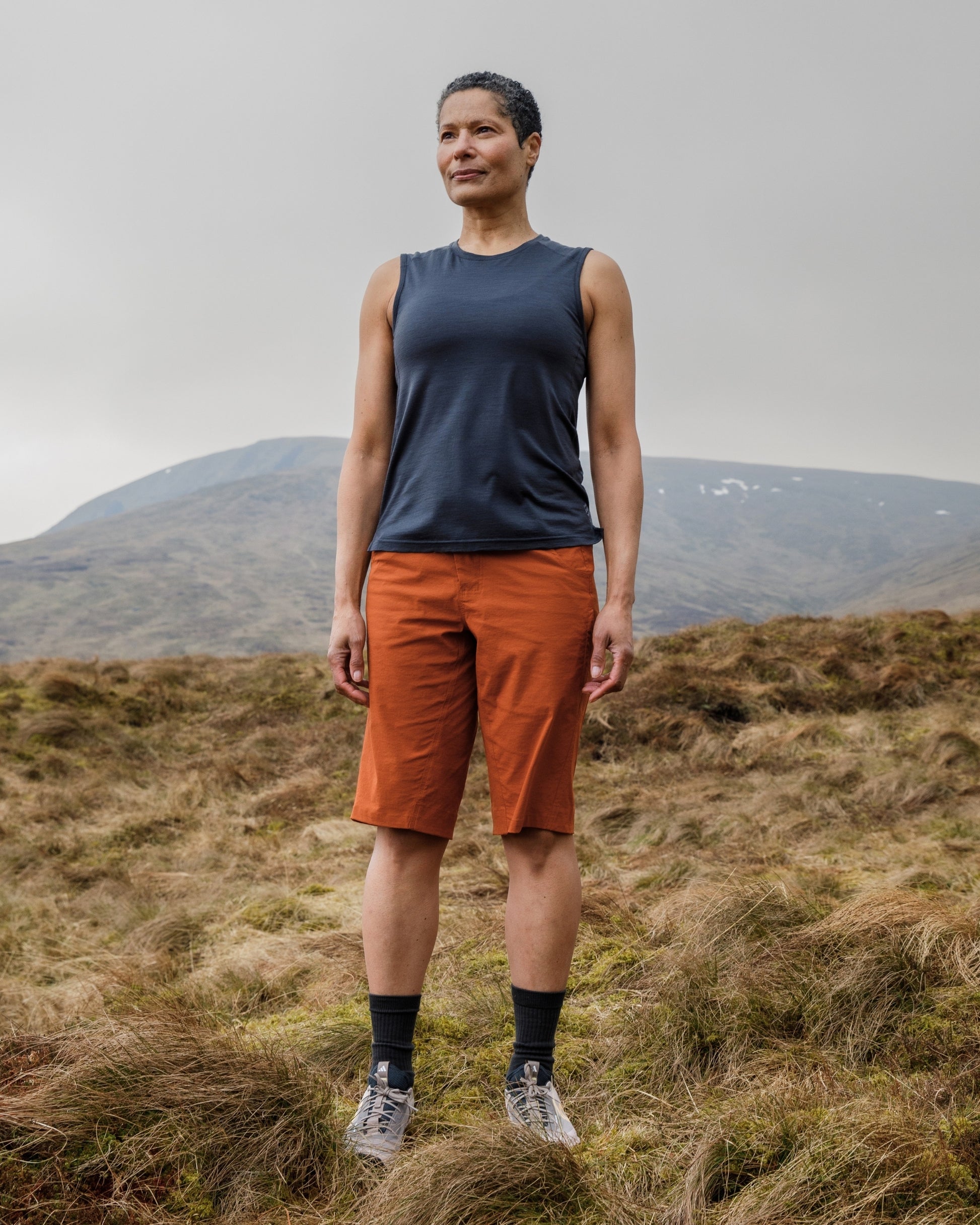 Person wearing a blue sleeveless shirt and orange shorts standing in a grassy field with mountains in the background.