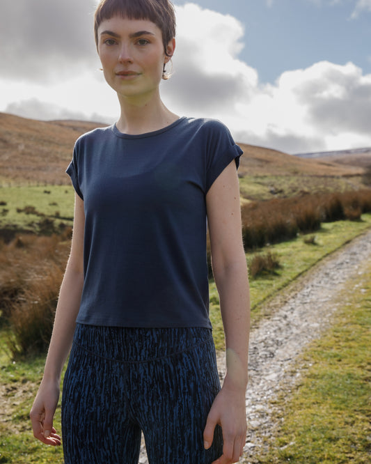 Woman standing on a path with a scenic landscape in the background wearing a blue merino t-shirt