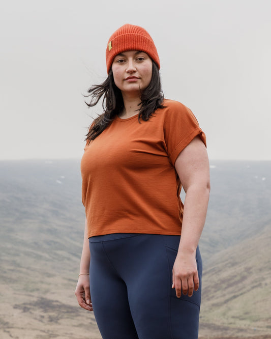 Person wearing a FINDRA orange Merino wool top and blue leggings standing in a field with mountains in the background