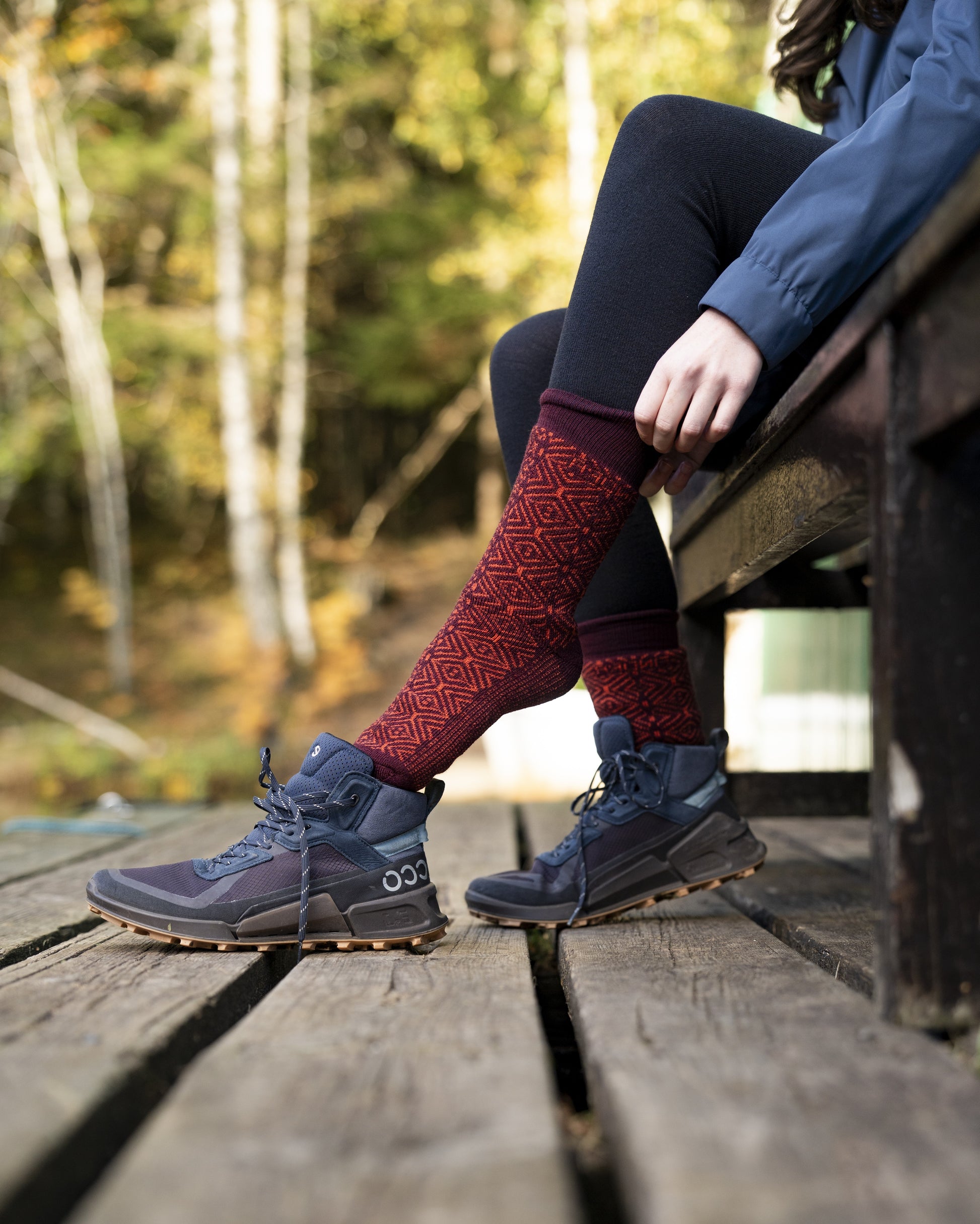 Person sitting on a wooden deck wearing red patterned socks and blue hiking boots with a forest background.