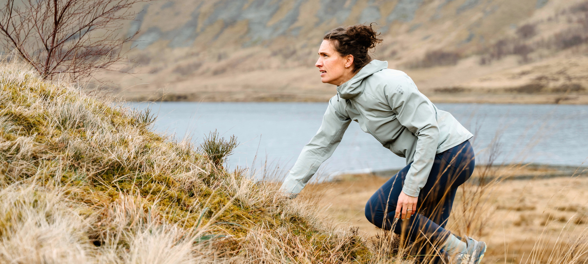 A woman in a light gray hooded jacket and dark blue leggings crouches in dry grass near a lake, with hills in the background.