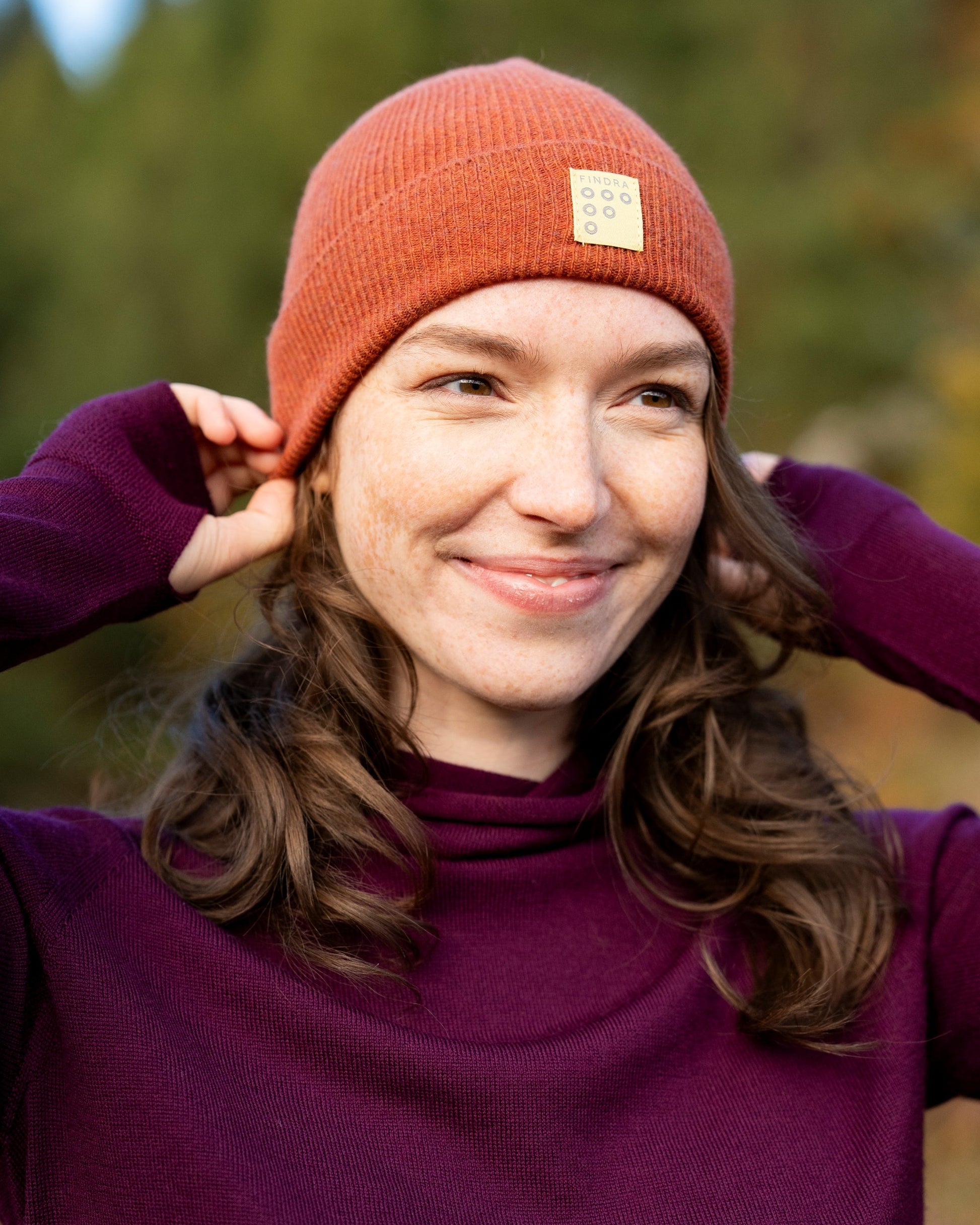 Person wearing a red beanie with a FINDRA logo, standing outdoors with trees in the background