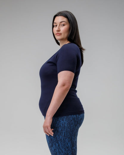 Woman wearing a navy blue top and jeans standing against a gray background