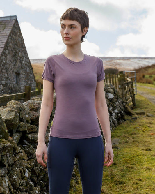 Woman in purple top and blue leggings standing in a rural setting with a stone wall and grassy field.