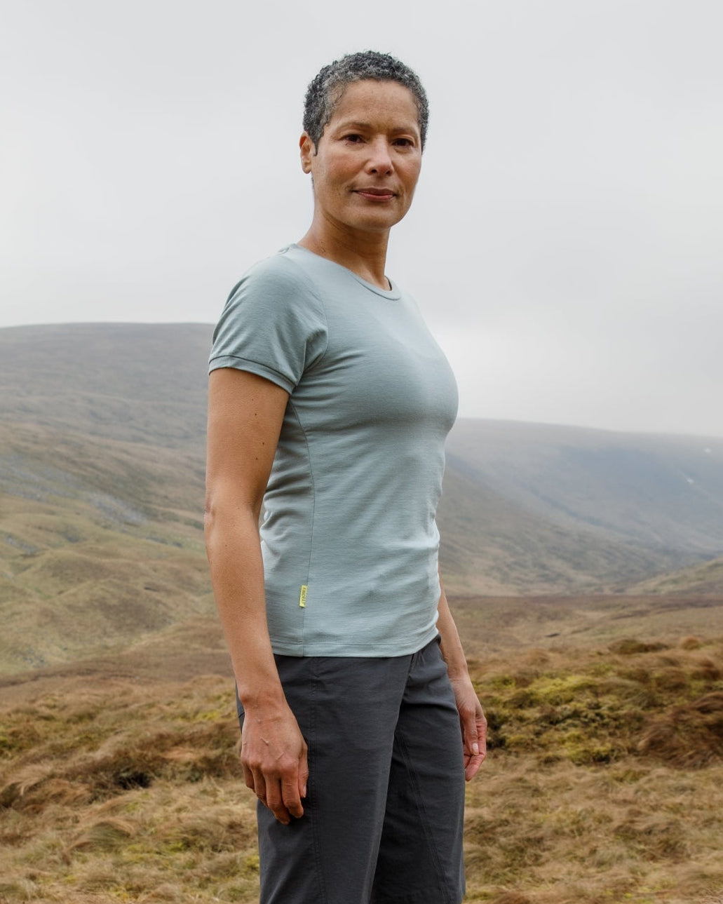 Person standing in a grassy field with mountains in the background wearing a Merino T-Shirt from FINDRA
