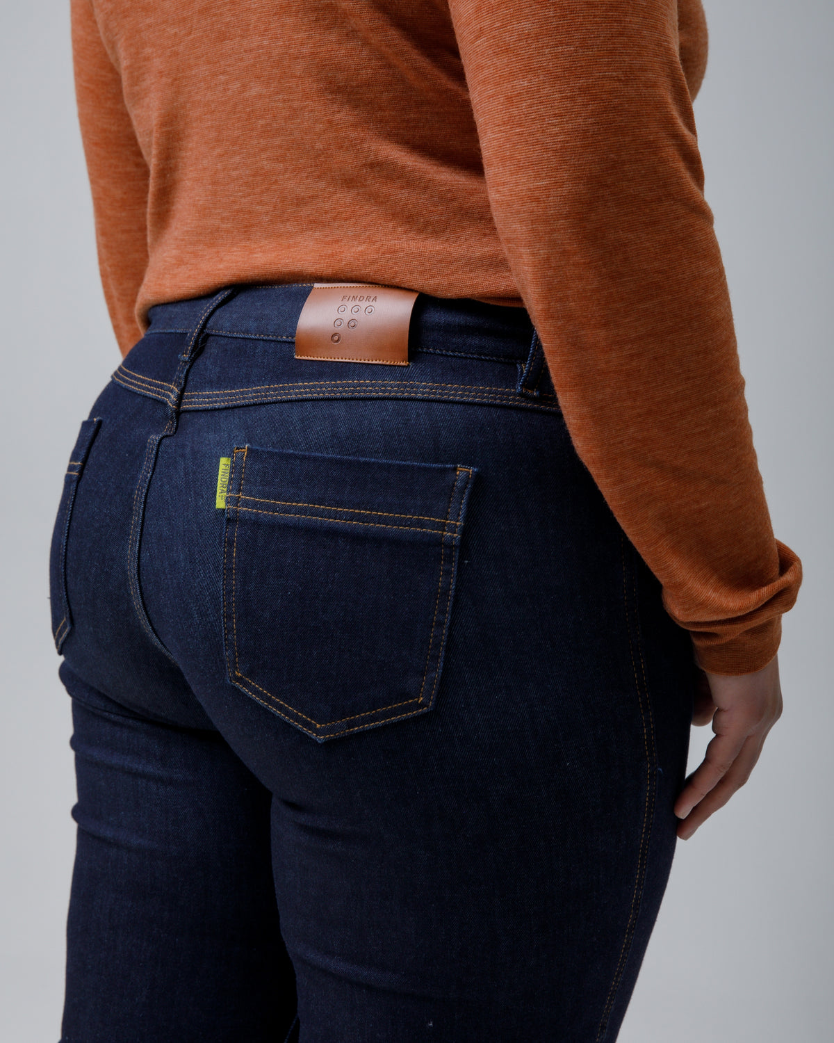 Rear view of a person wearing dark indigo blue jeans with a brown leather waistband patch and an orange-brown long-sleeve top, against a solid gray studio background.