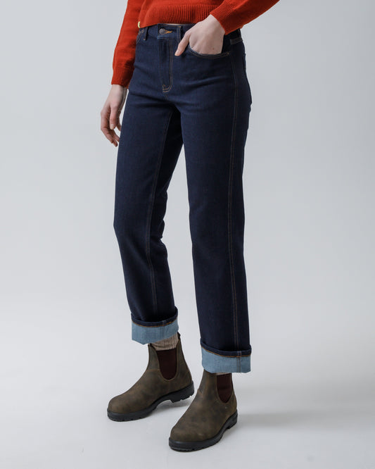 A model stands against a light gray studio background wearing a bright red long-sleeve knit sweater, dark blue jeans with rolled cuffs, and brown Chelsea-style boots.