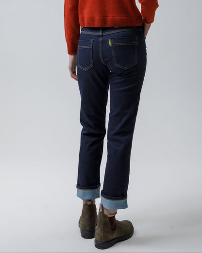 A model shown from the back wearing a rust-orange long-sleeve knit sweater, dark blue jeans with cuffed hems, and brown suede boots, set against a light gray studio background.